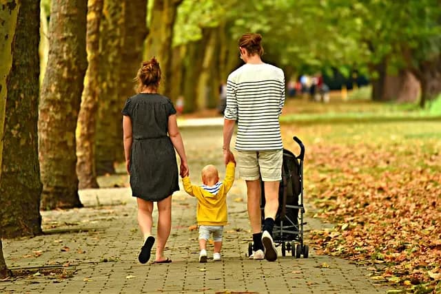 Two parents walking a toddler between them down a tree-lined path in autumn